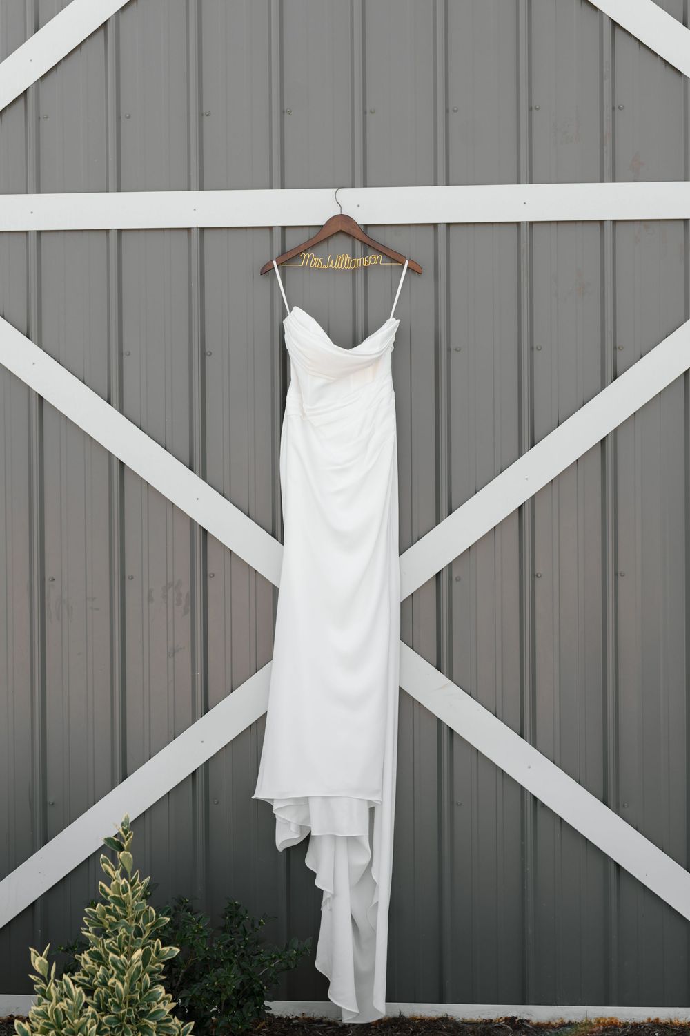 White spaghetti strap wedding gown hanging on wooden hanger against gray barn door with white trim.