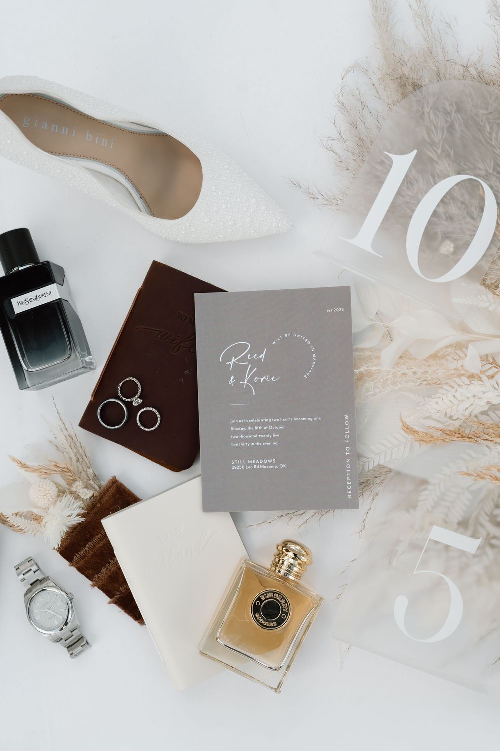 Elegant wedding flat lay with bridal heels, perfume, rings, watch, and gray invitation on white background.