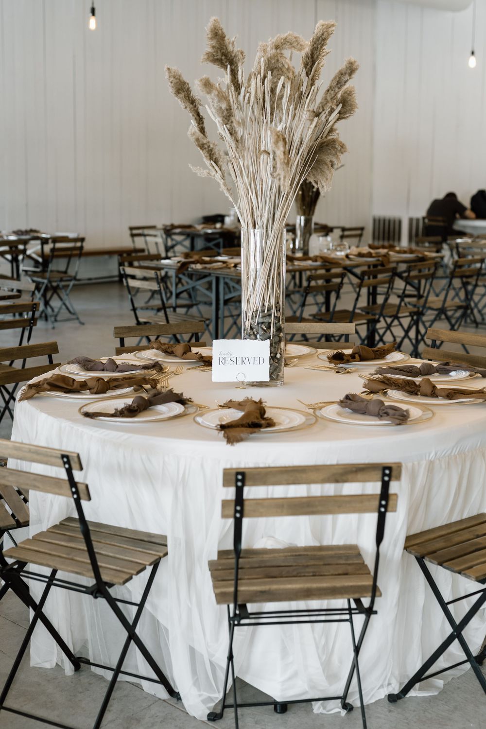 Elegant wedding reception table with white linen, pampas grass centerpiece, wooden chargers, and folding chairs in a bright venue.