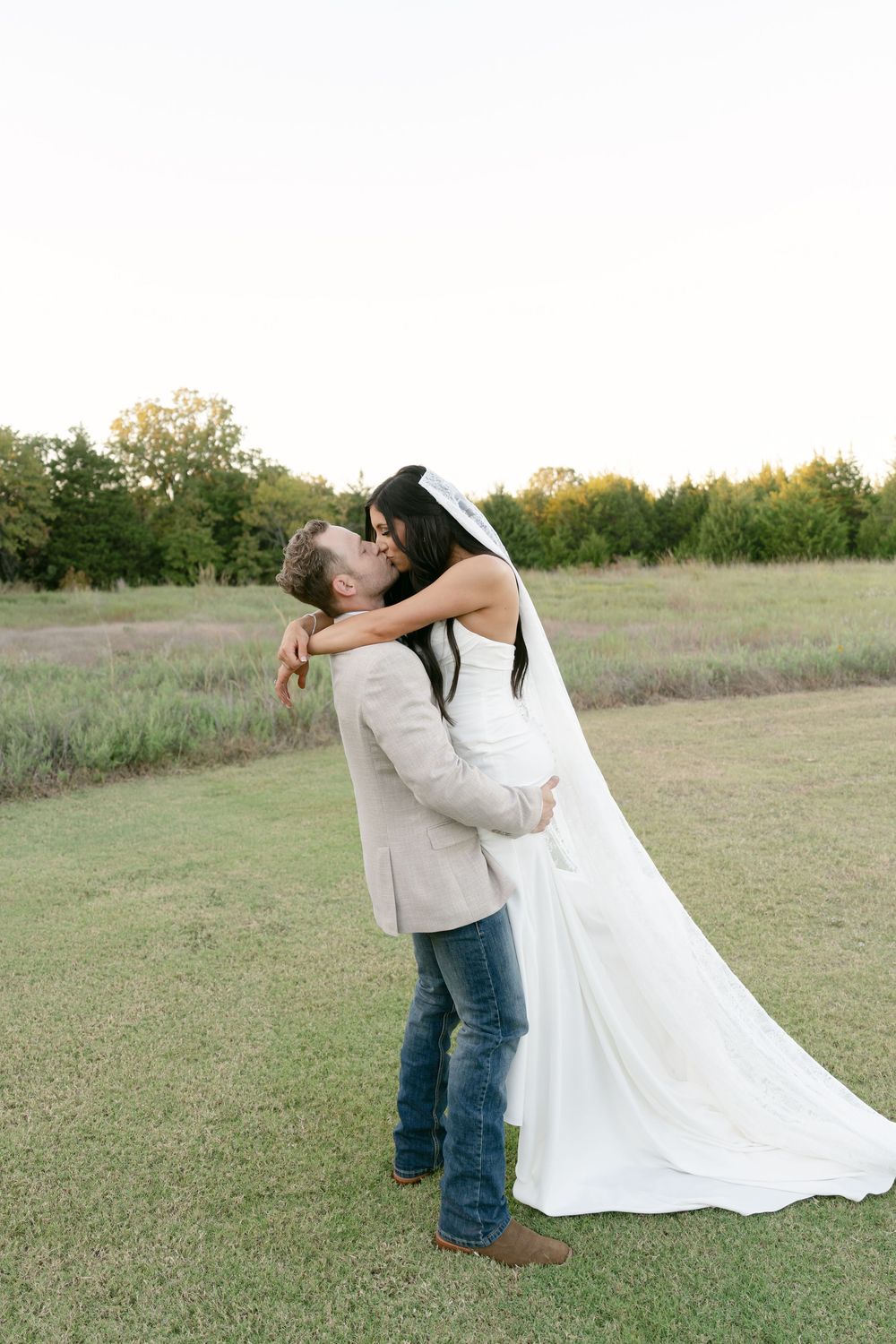 Bride and groom sharing a romantic kiss in an open field at golden hour during their wedding.