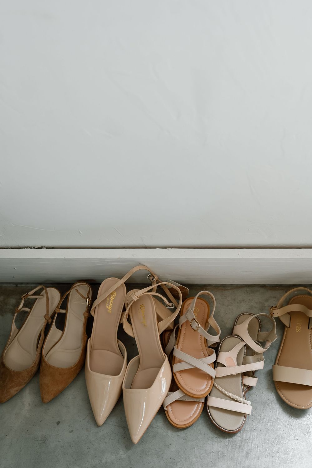 Four pairs of nude beige women's heels and sandals neatly arranged on a gray floor against a white wall.