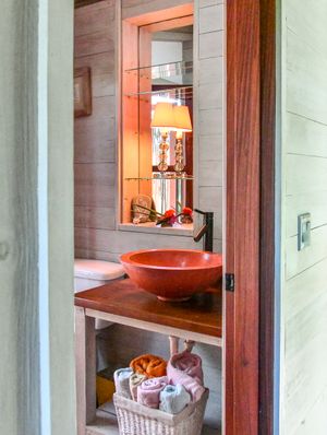 Rustic bathroom with a terracotta vessel sink on a wooden vanity, mirror, warm lamp, and neatly stored towels in a wicker basket.