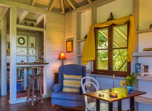 Cozy tropical cottage interior with blue armchair, wooden walls, yellow curtains, and a bar area visible through an open doorway.