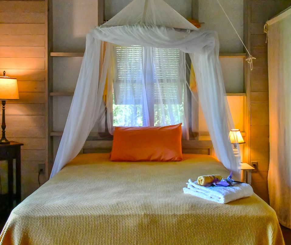 Cozy canopy bed with sheer white drapes, orange pillow, yellow bedspread, and folded towels in a rustic wooden bedroom.