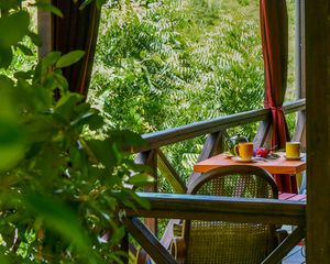 Cozy balcony with wicker chair, orange table, and mugs overlooking lush green tropical garden.