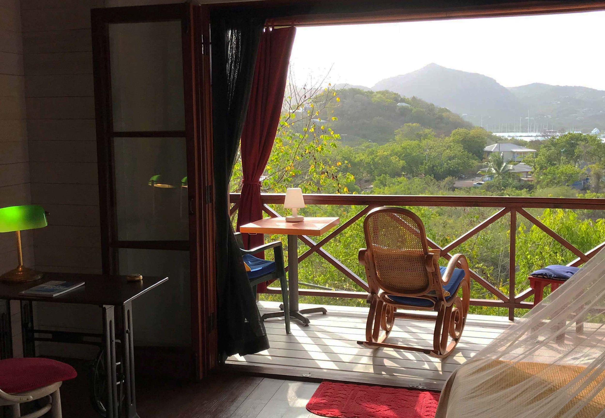 Cozy hotel room balcony with rattan chairs, red curtains, and lush mountain views in a tropical setting.
