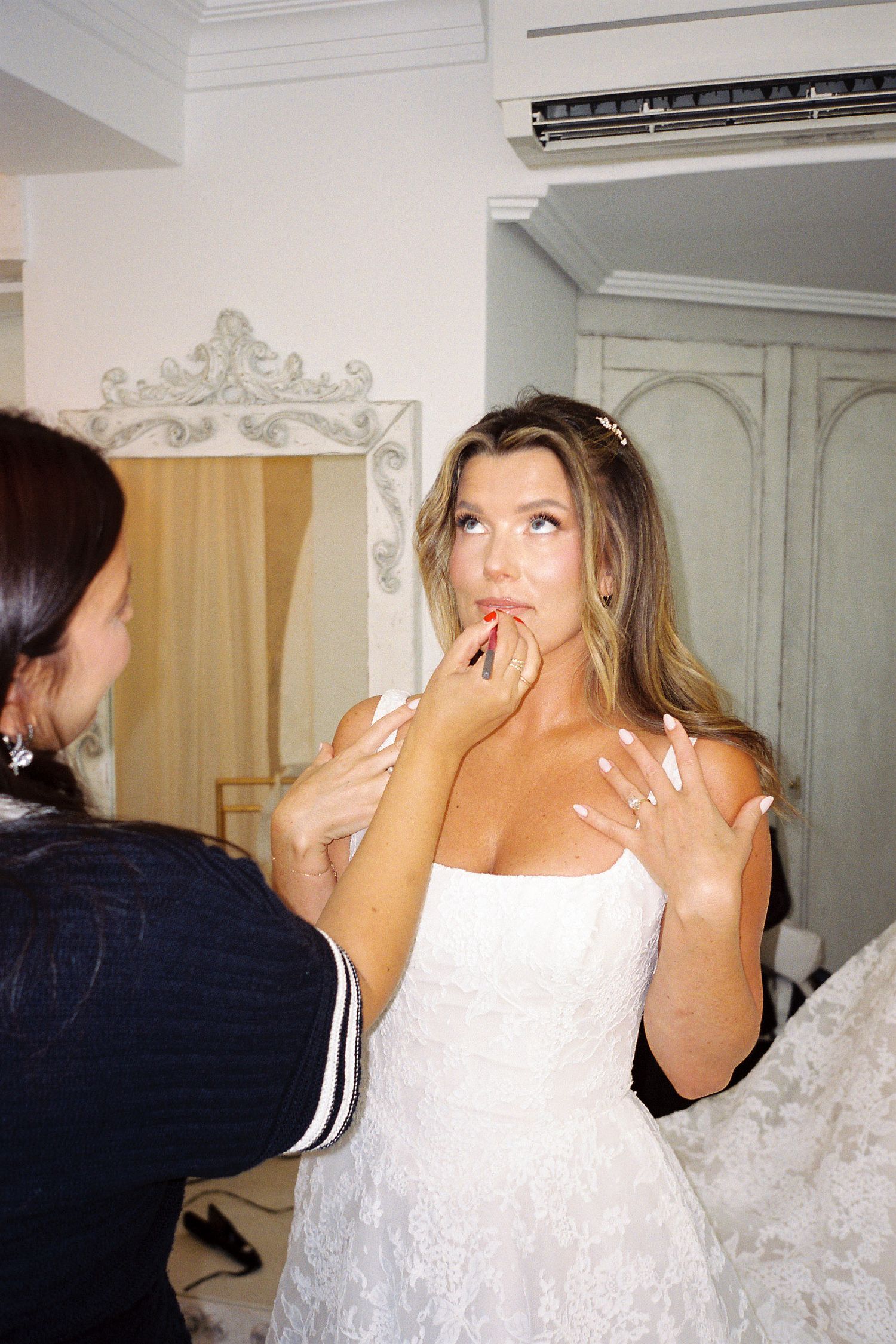 Makeup artist applying lipstick to a bride in a white strapless wedding dress during bridal preparations.
