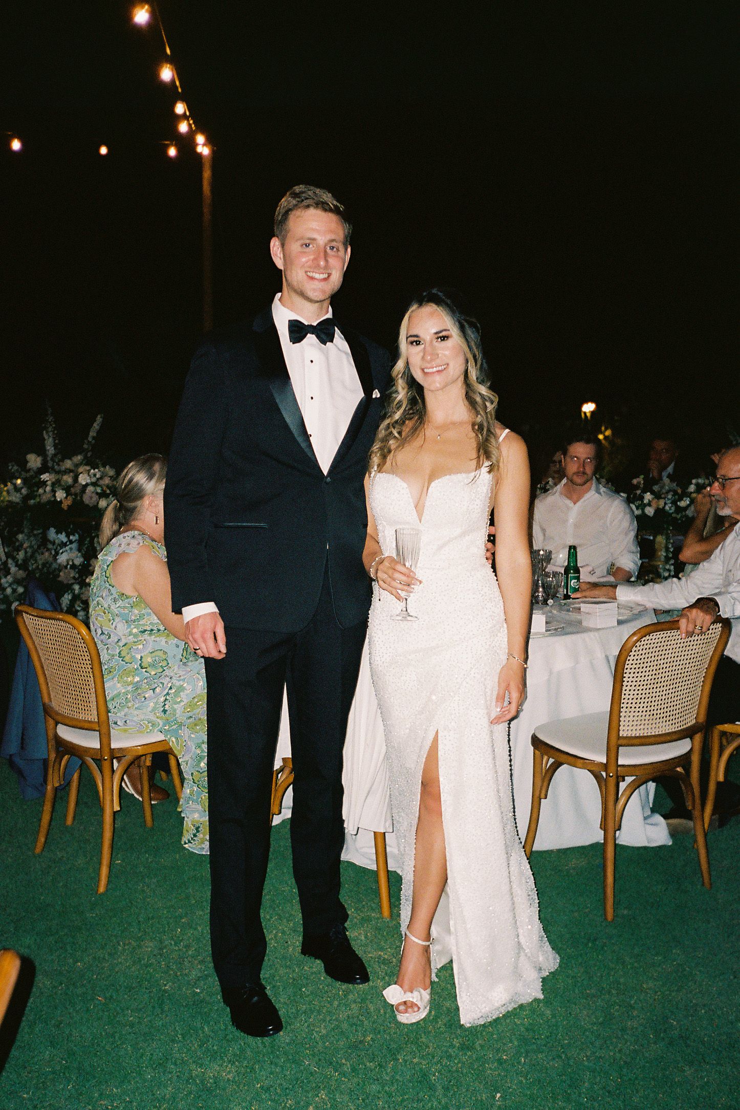 Couple in formal wedding attire, groom in black tuxedo and bride in white gown, posing at outdoor evening reception.