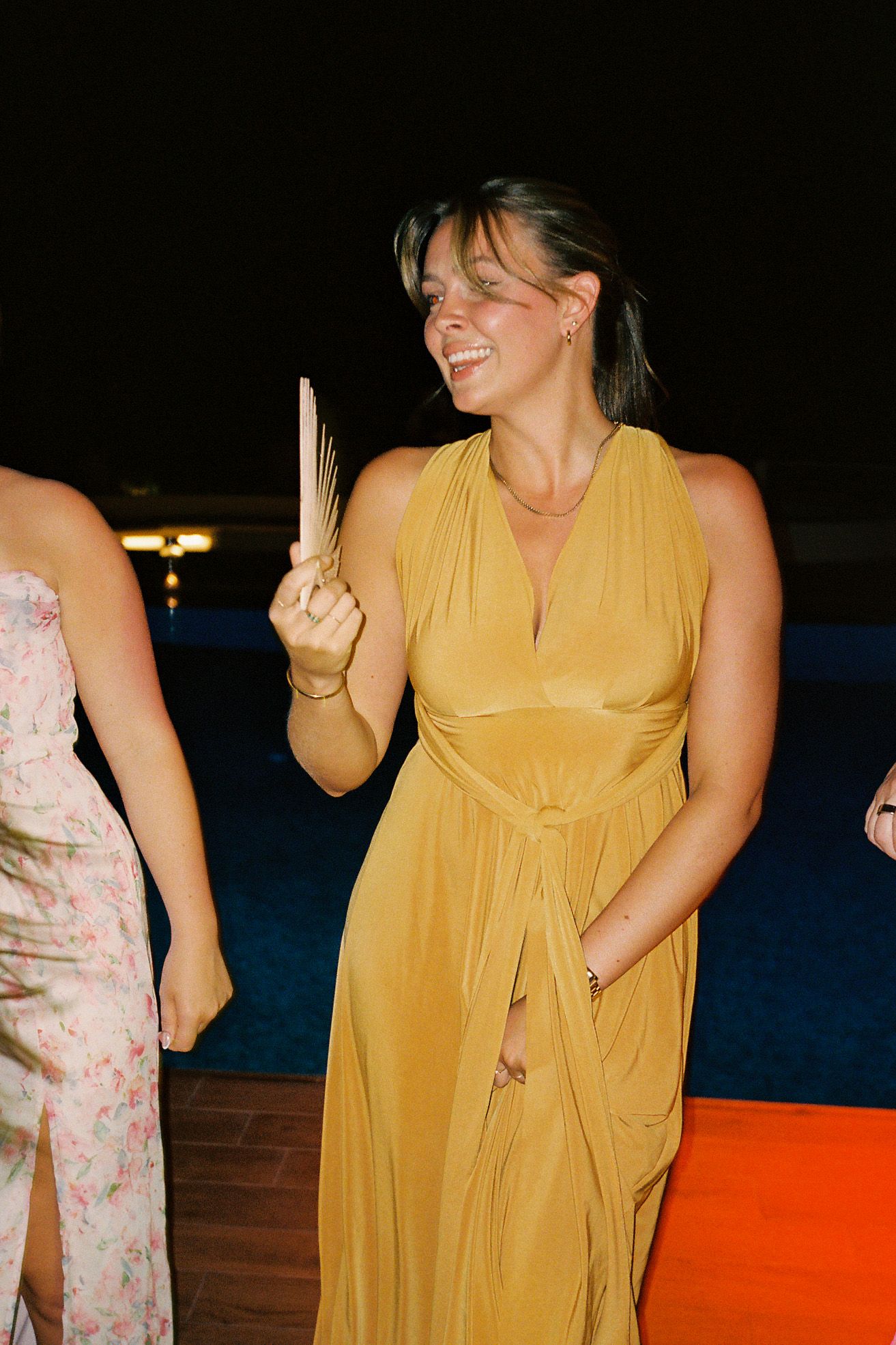 Woman in yellow halter dress holding a fan, laughing at an outdoor evening event.