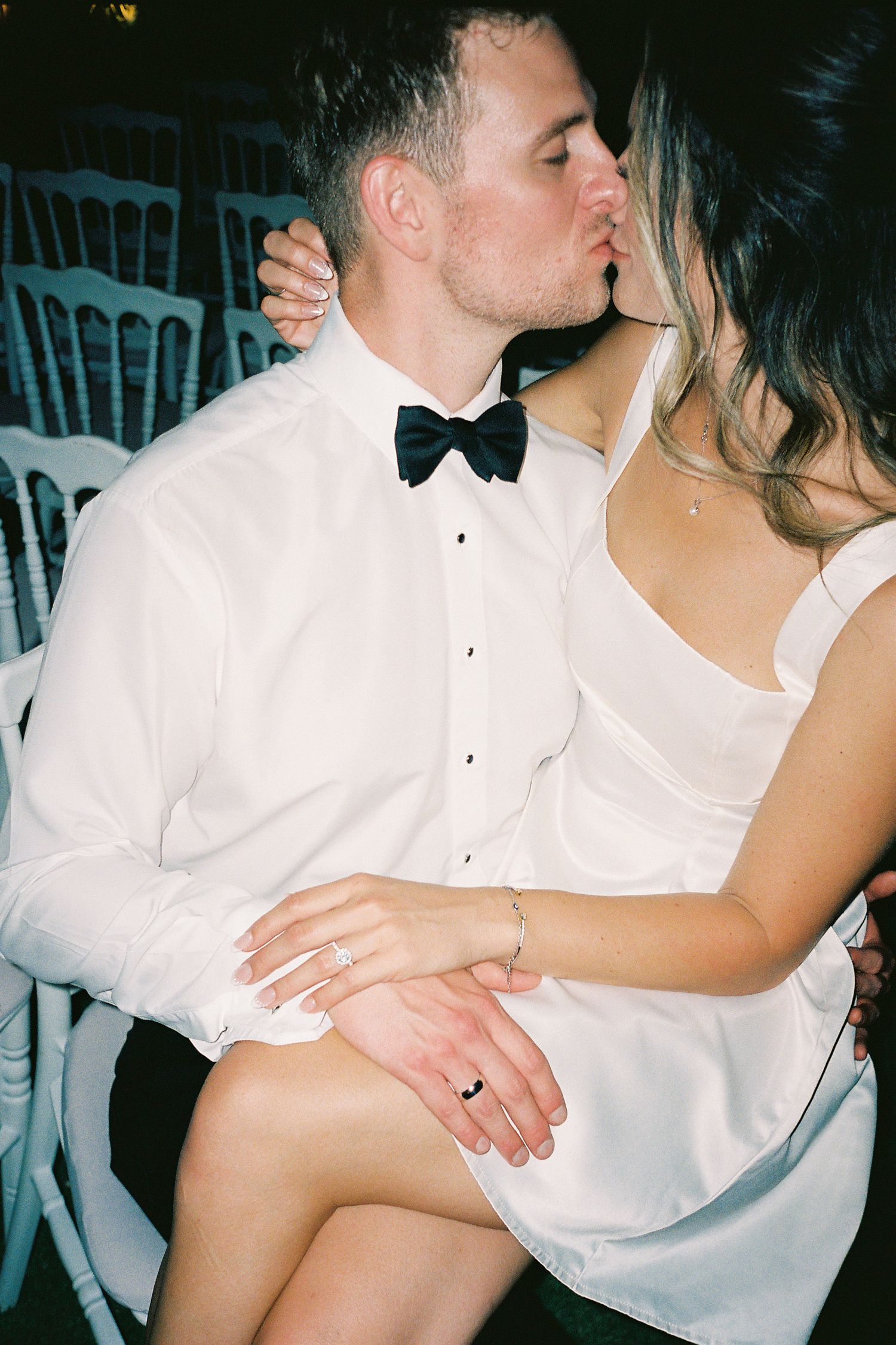 Couple in wedding attire sharing a kiss, groom in white shirt and bow tie, bride in white dress with rings visible.