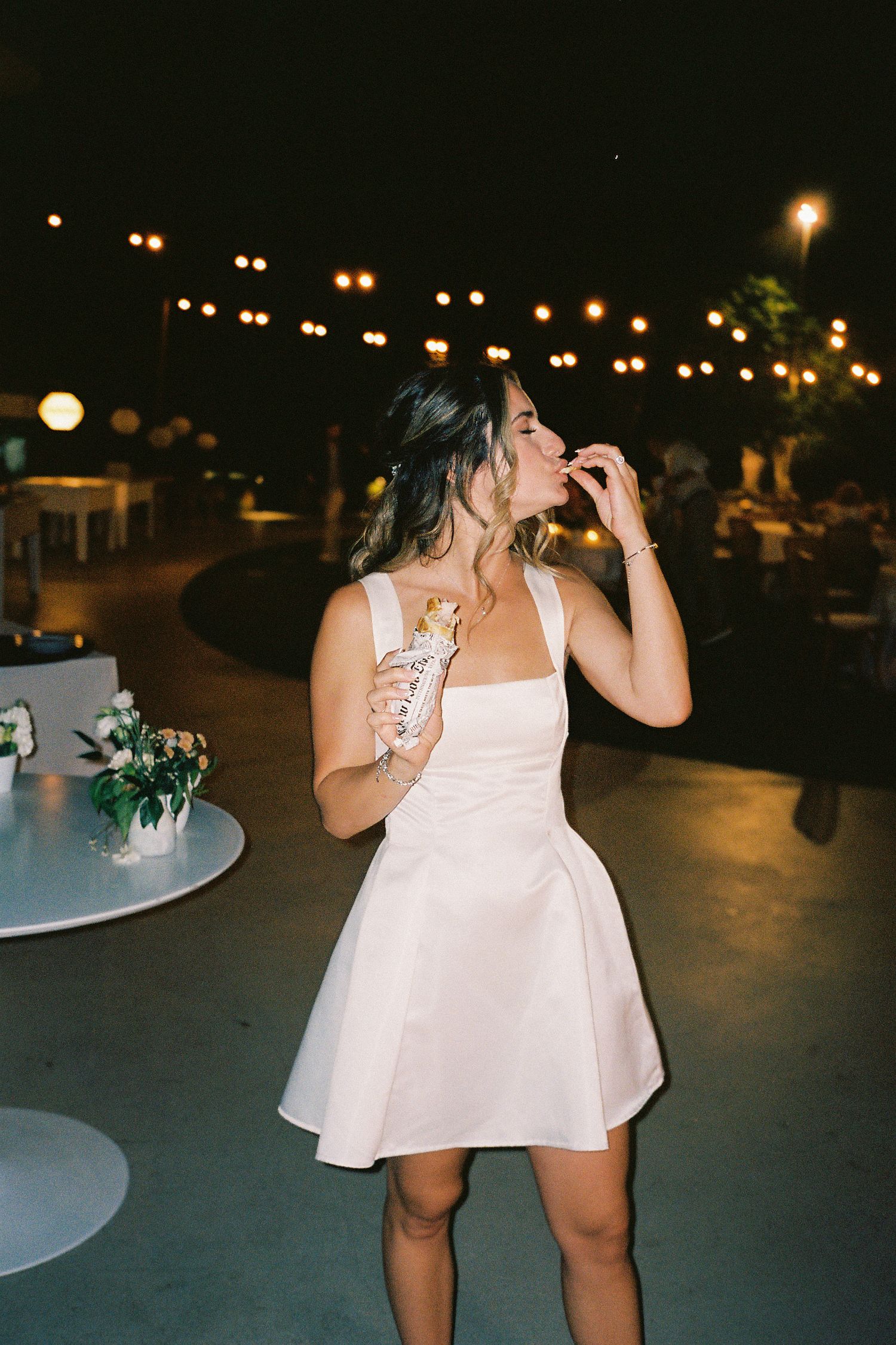 Woman in white mini dress holding champagne at outdoor evening wedding reception with string lights.