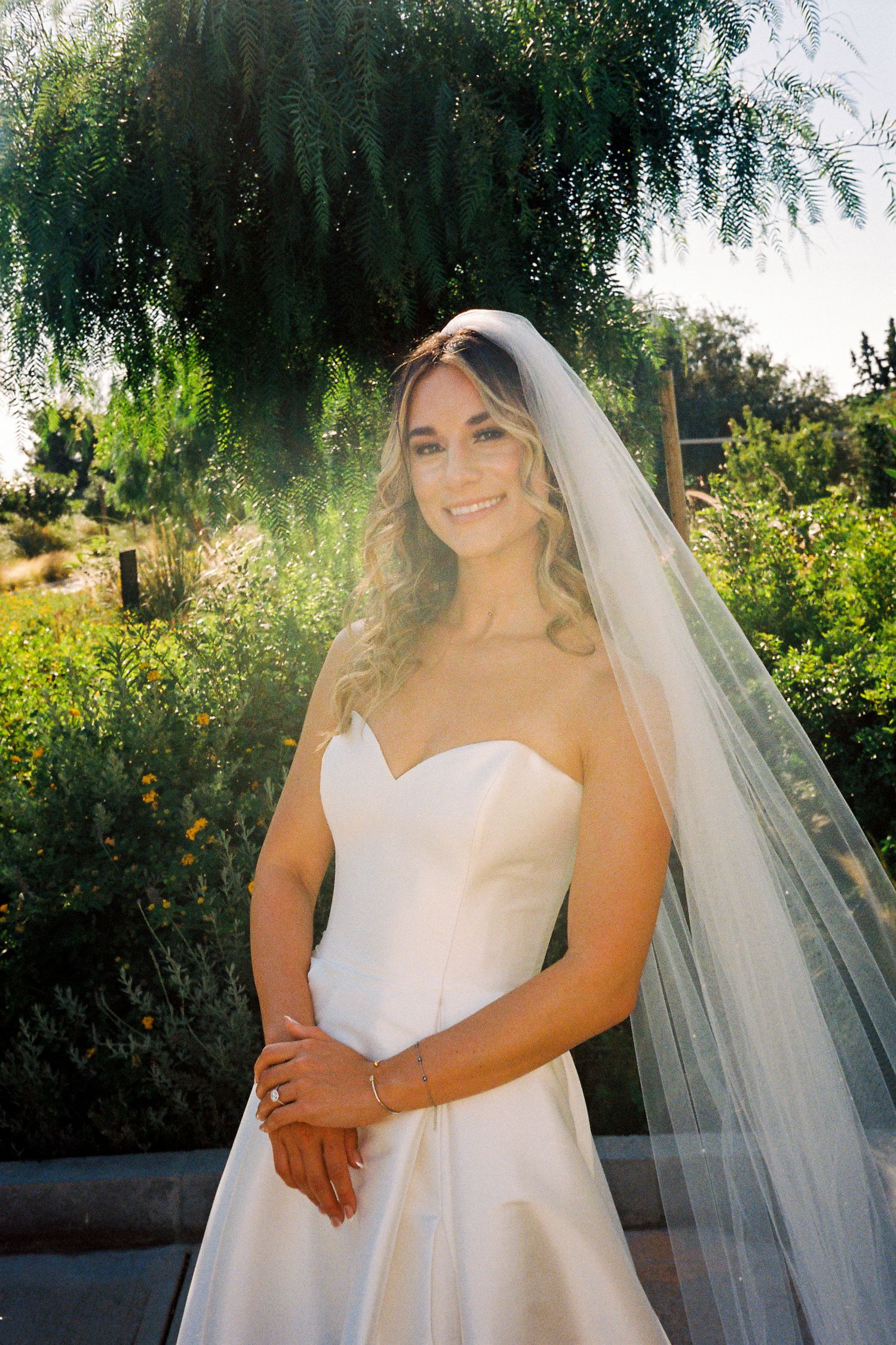 Smiling bride in strapless white gown and flowing veil poses outdoors in lush green garden with warm sunlight.