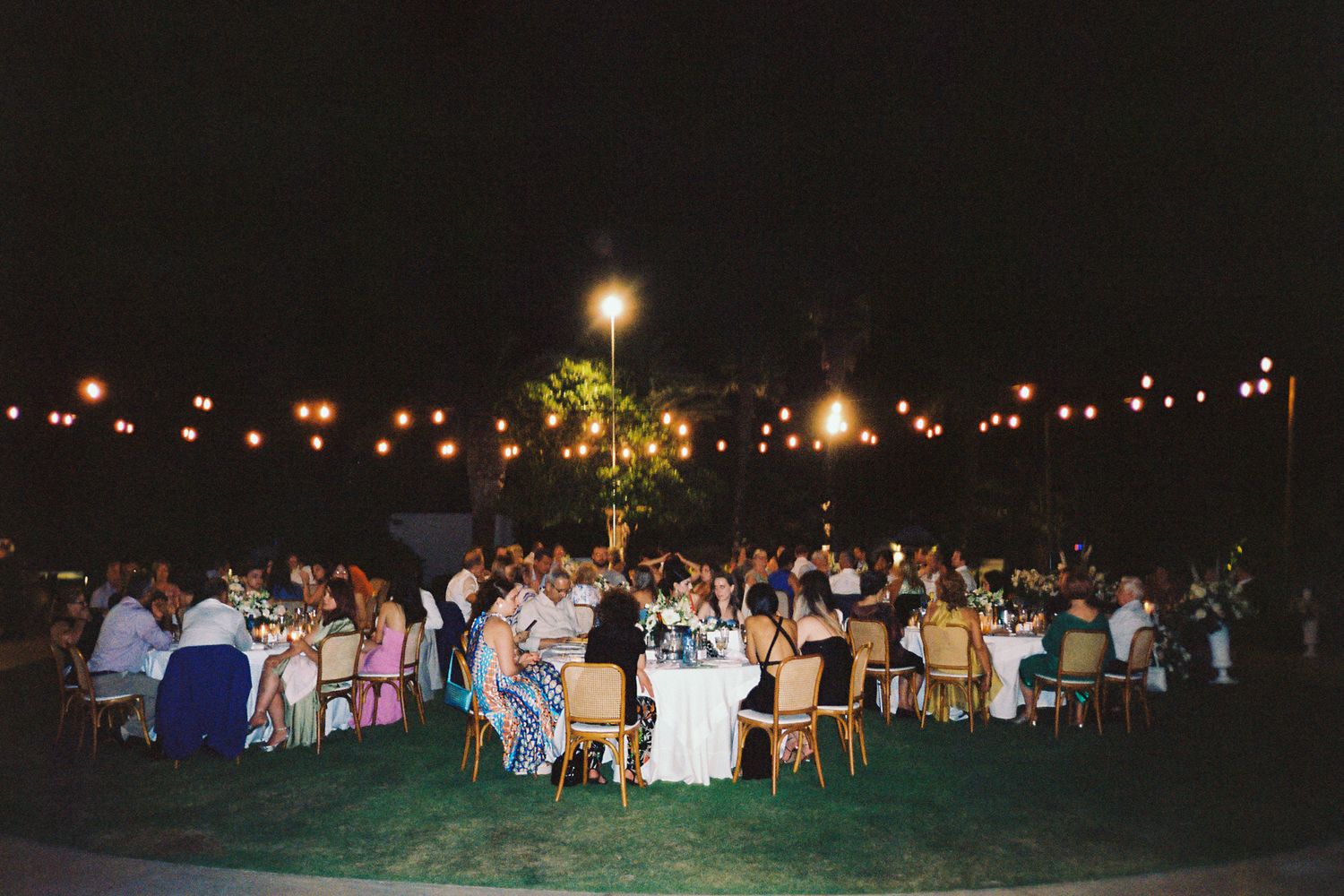 Outdoor evening wedding reception with string lights, guests seated at round tables on green lawn under night sky.