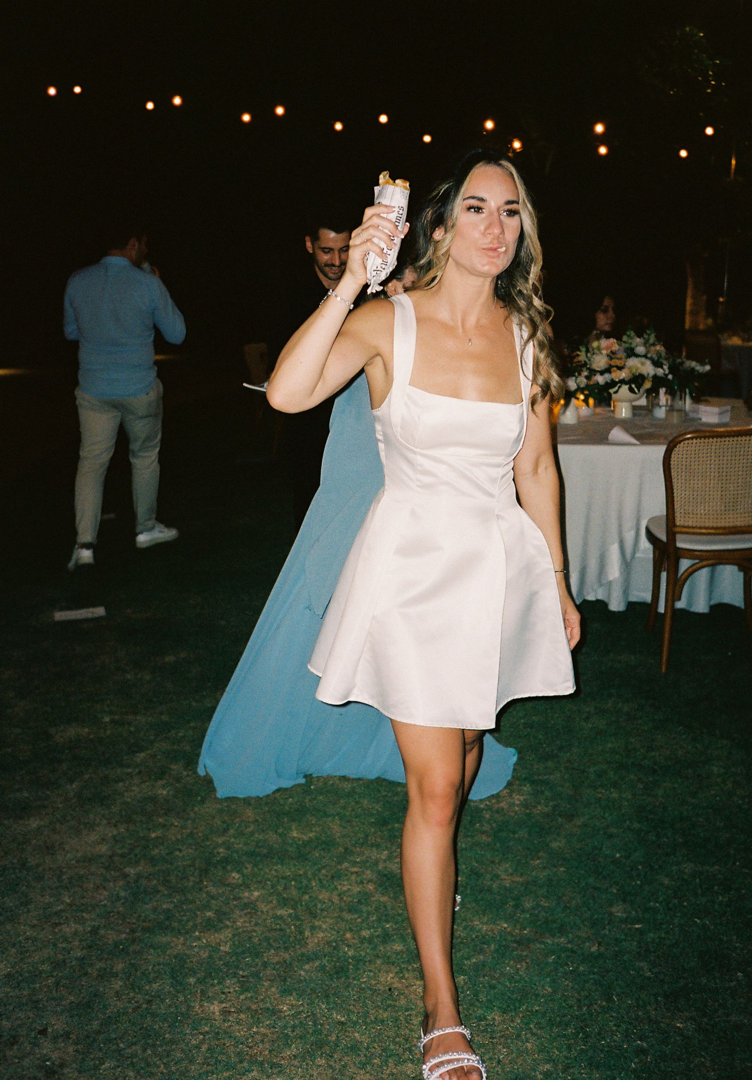 Woman in white mini dress holding a drink at an outdoor evening wedding reception with string lights.