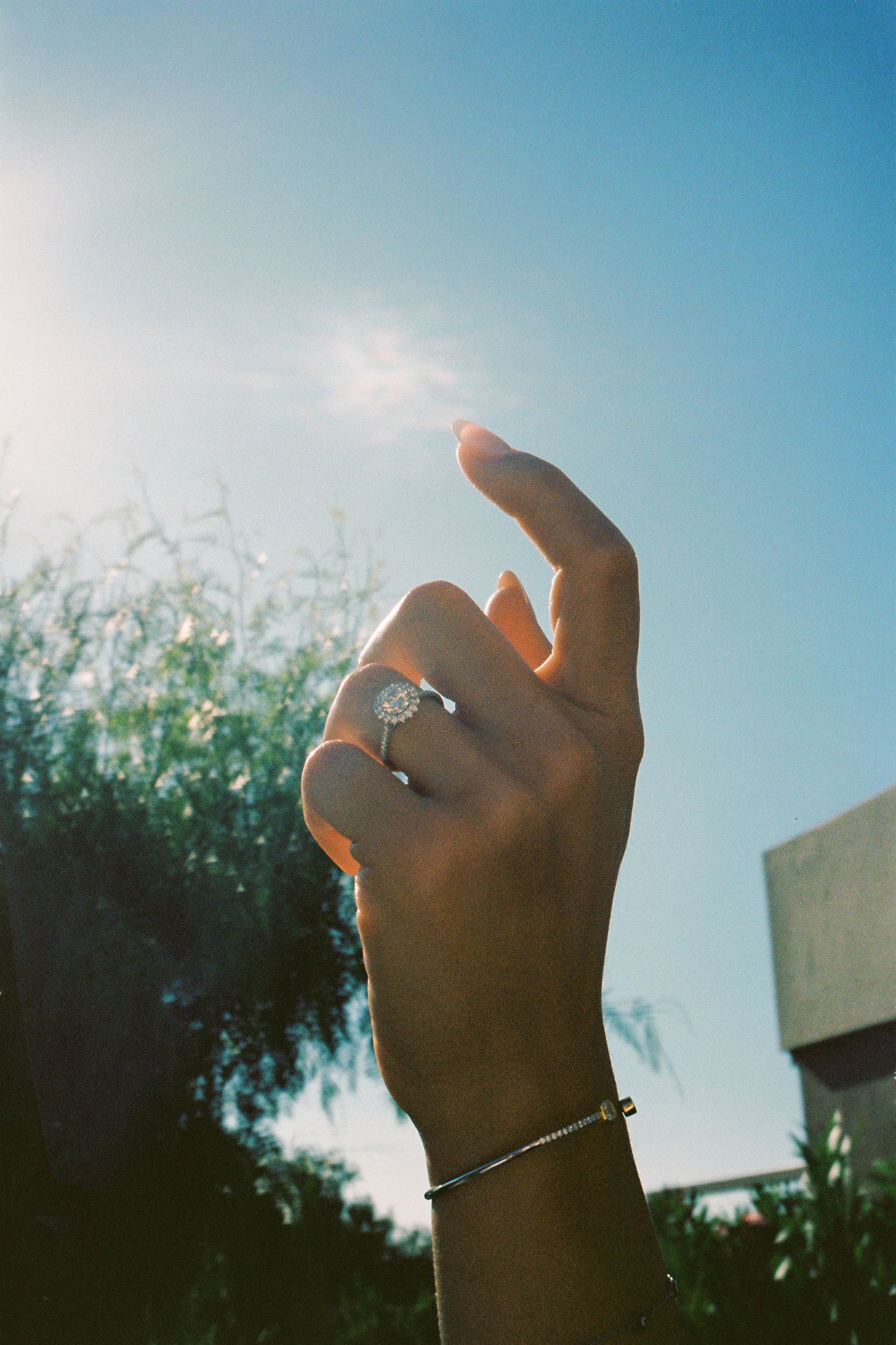 Hand raised toward sun wearing diamond halo engagement ring and delicate bracelet against blue sky.