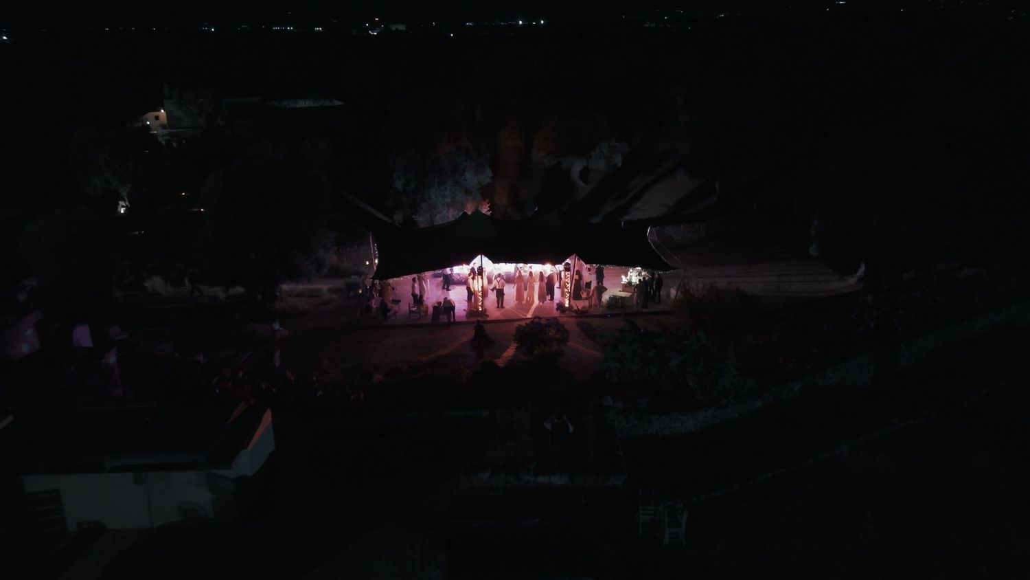 Aerial night view of an illuminated outdoor stage performance with colorful lights surrounded by darkness.