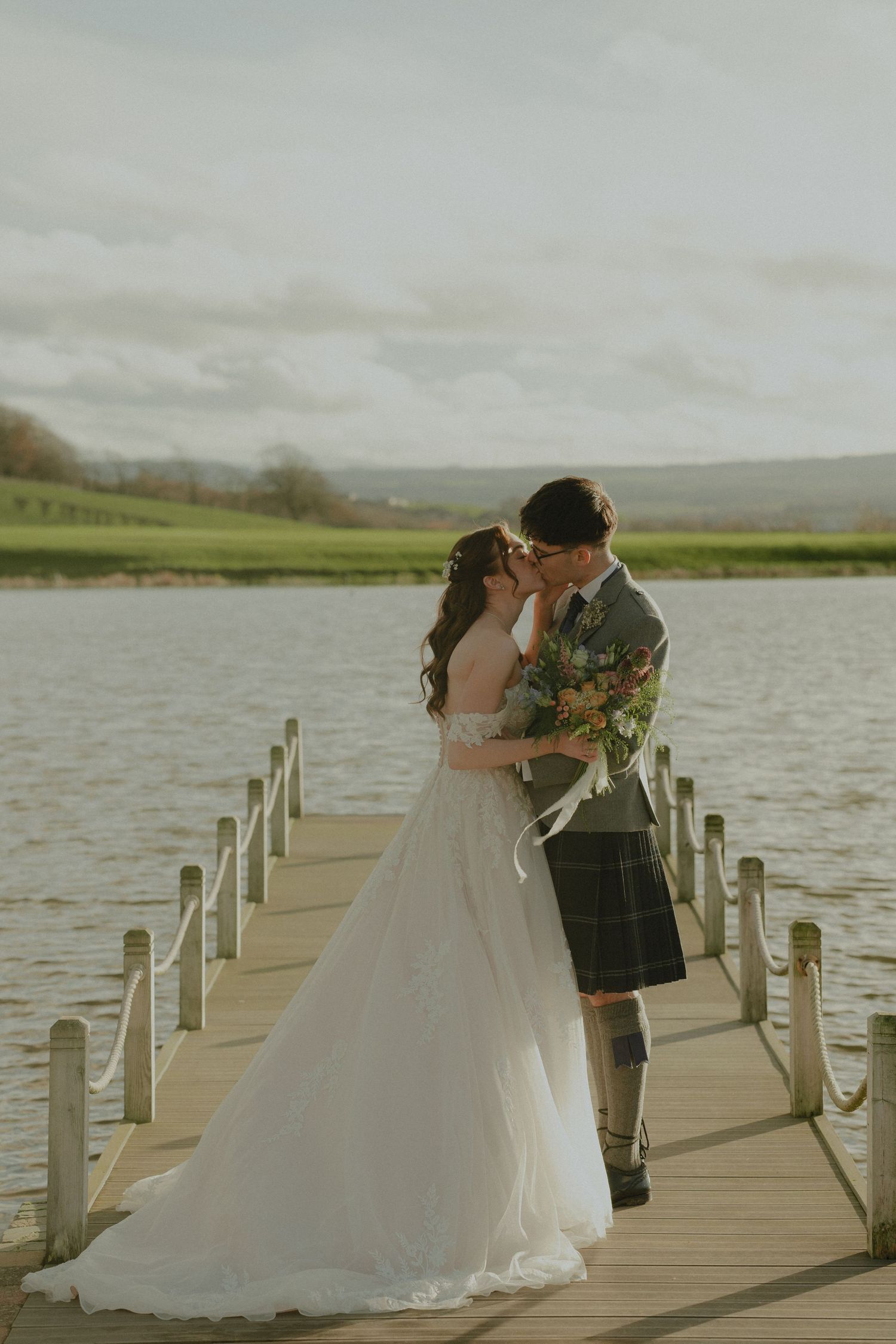 Bride and groom share a kiss on a wooden dock by a loch, wearing wedding dress and Scottish kilt.