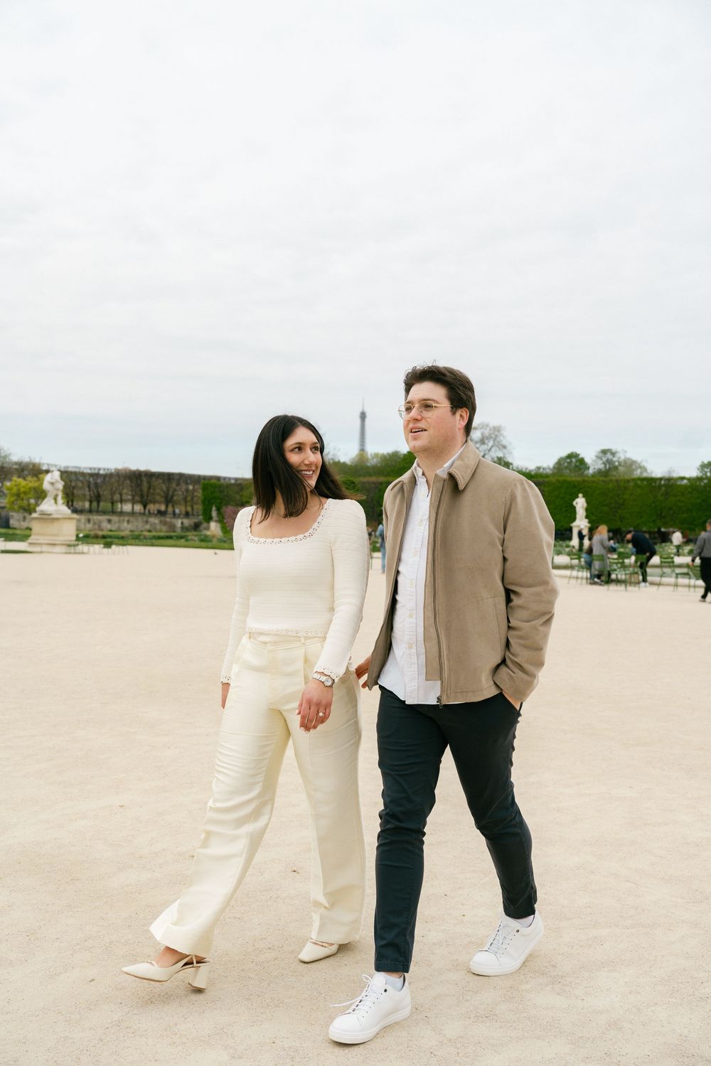 Couple walking hand in hand in Tuileries Garden, Paris, with the Eiffel Tower visible in the background.