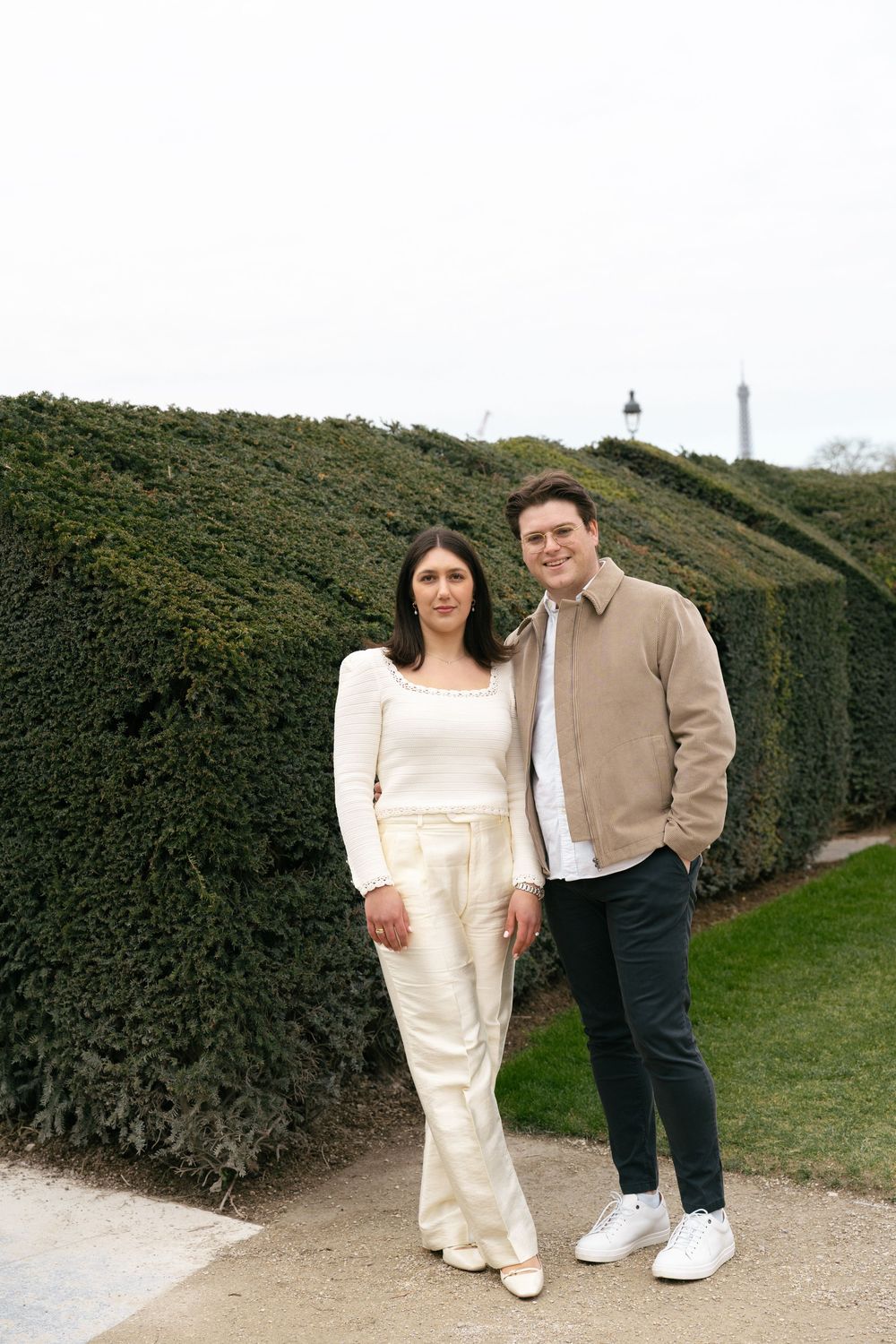Couple posing together in front of a tall hedge in a Parisian garden with the Eiffel Tower visible.