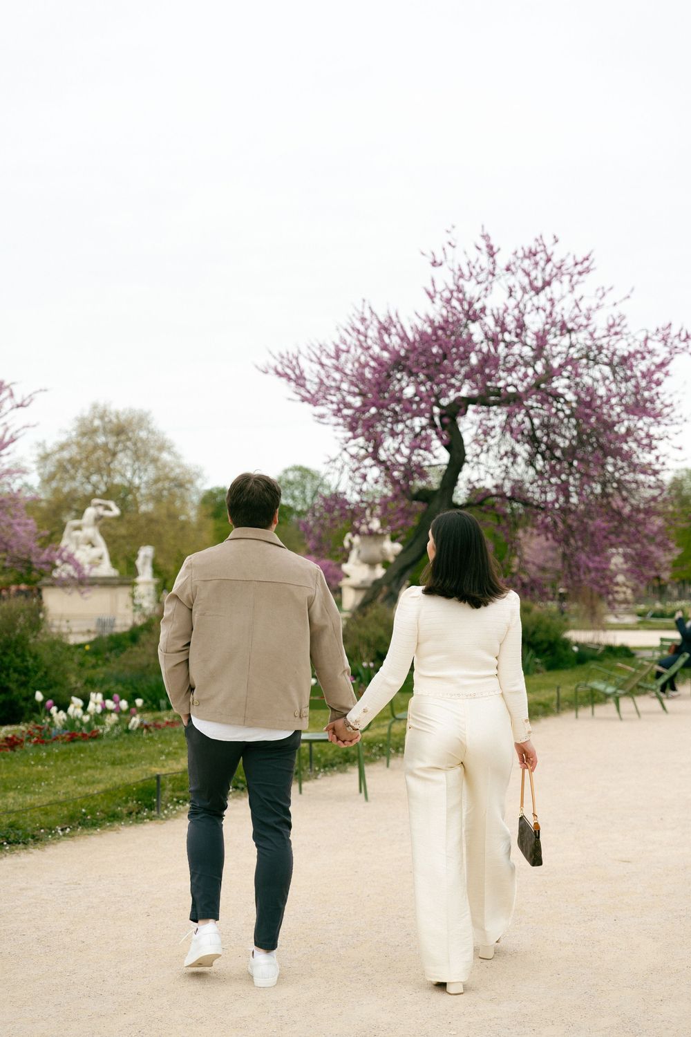 Couple holding hands walking through a Paris garden with pink cherry blossom trees in spring.