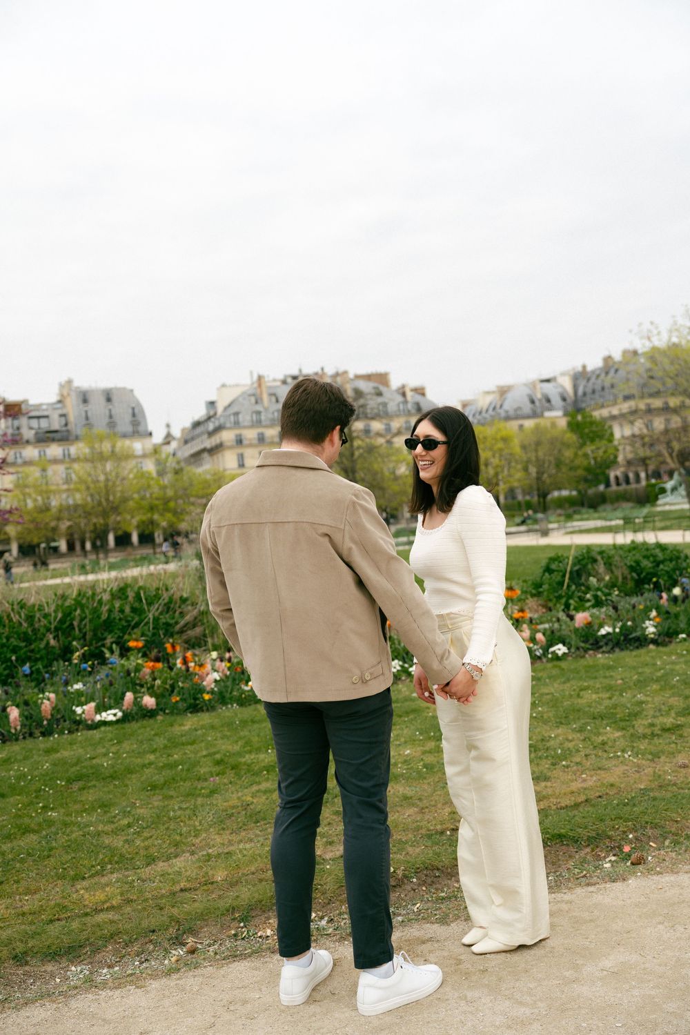 Couple holding hands in a Paris park, woman in white outfit with sunglasses, man in beige jacket, Haussmann buildings behind.
