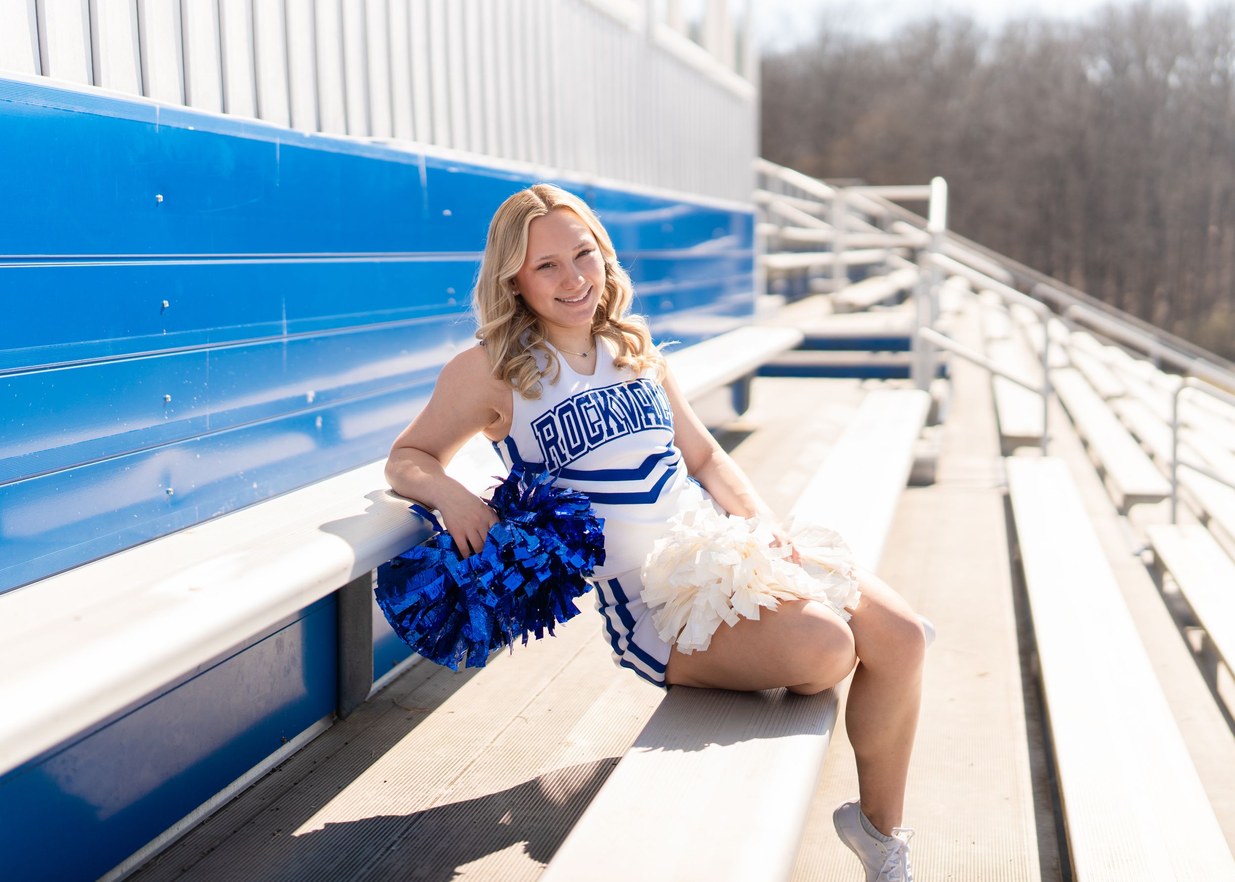 Cheerleader in blue and white uniform sitting on bleachers holding blue and white pom-poms on a sunny day.