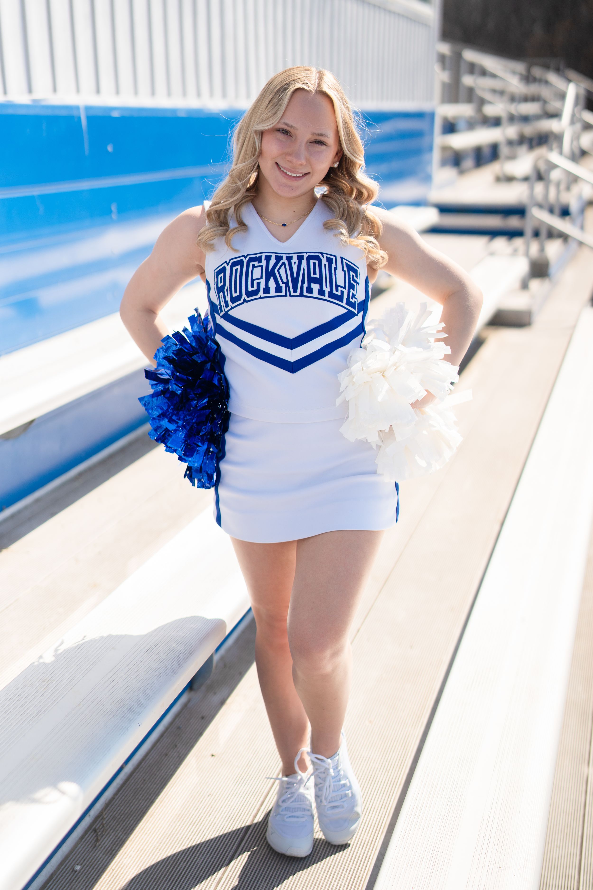 Rockvale cheerleader in white and blue uniform holding blue and white pom poms while posing in stadium bleachers.
