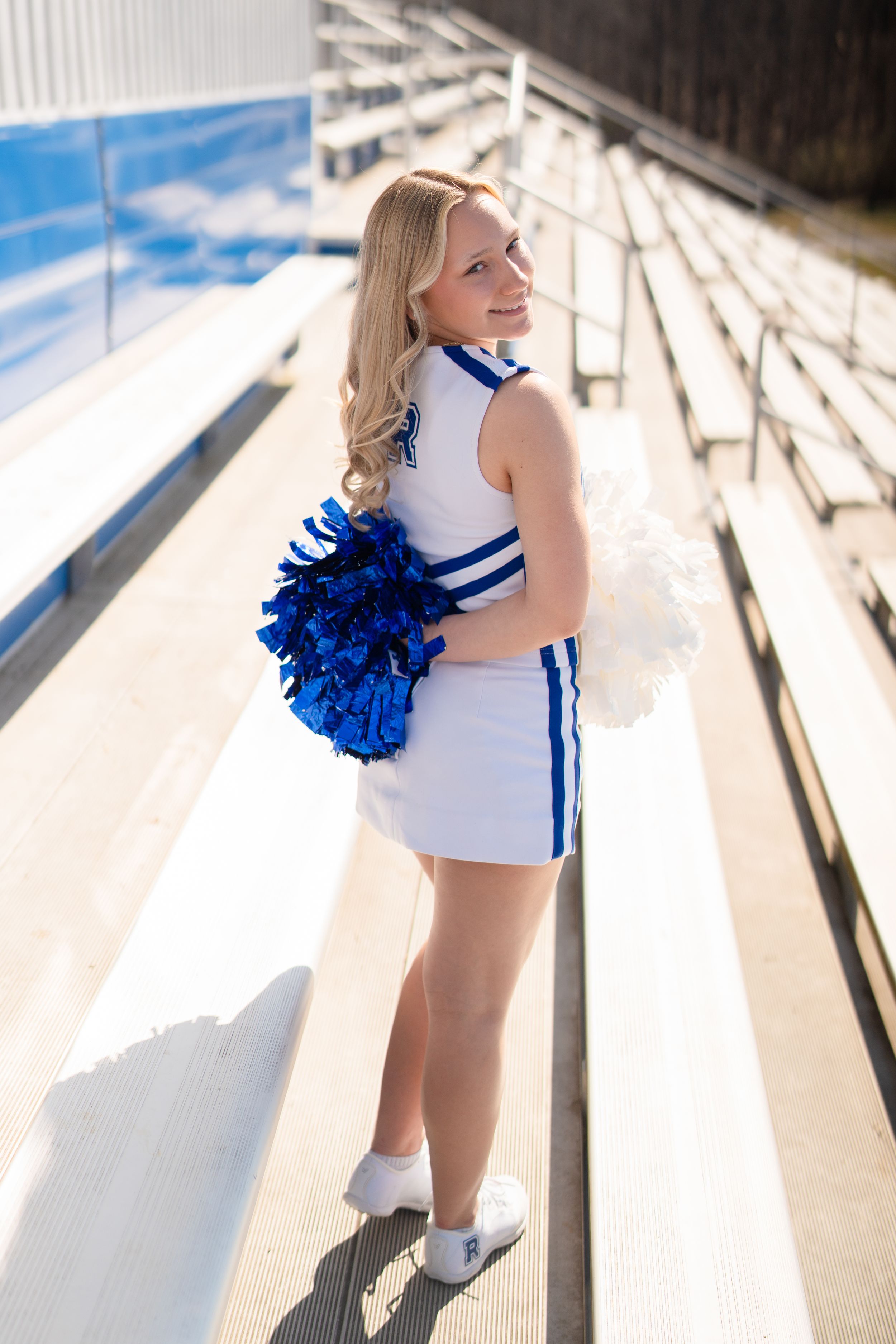 Cheerleader in white and blue uniform holding pom-poms stands on bleachers outdoors.