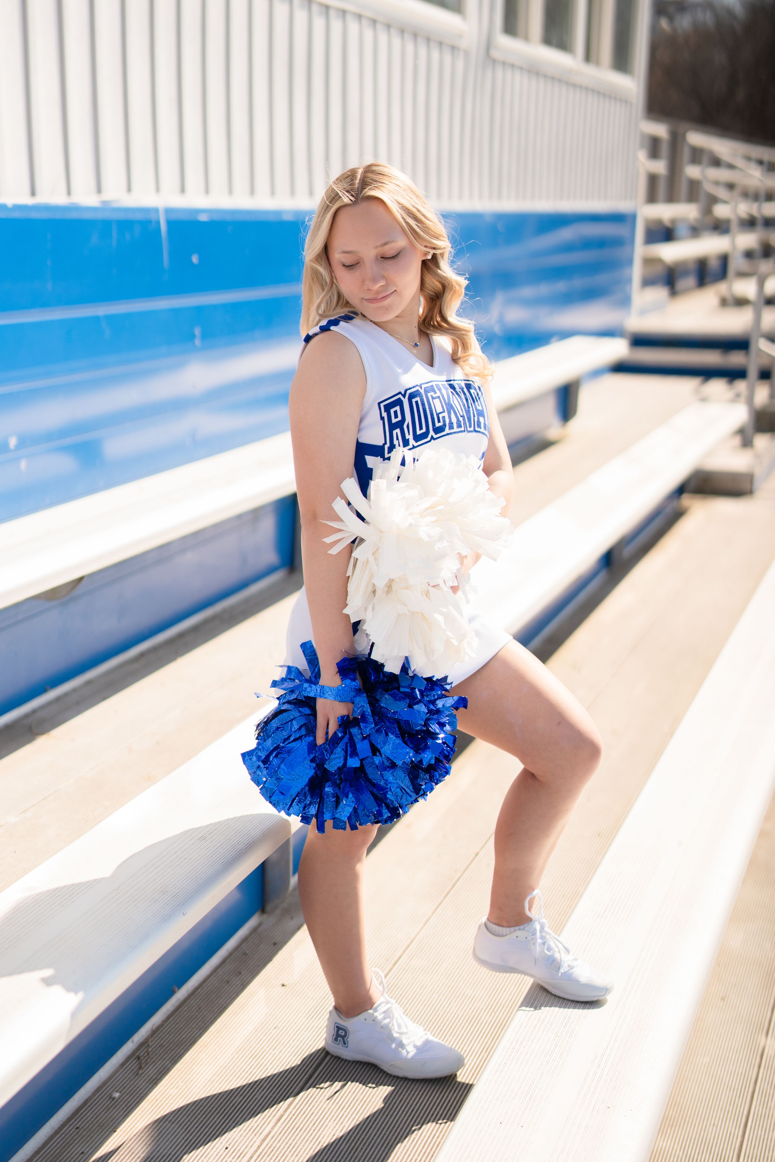 Rockvale cheerleader posing on stadium bleachers holding blue and white pom poms in her school uniform.