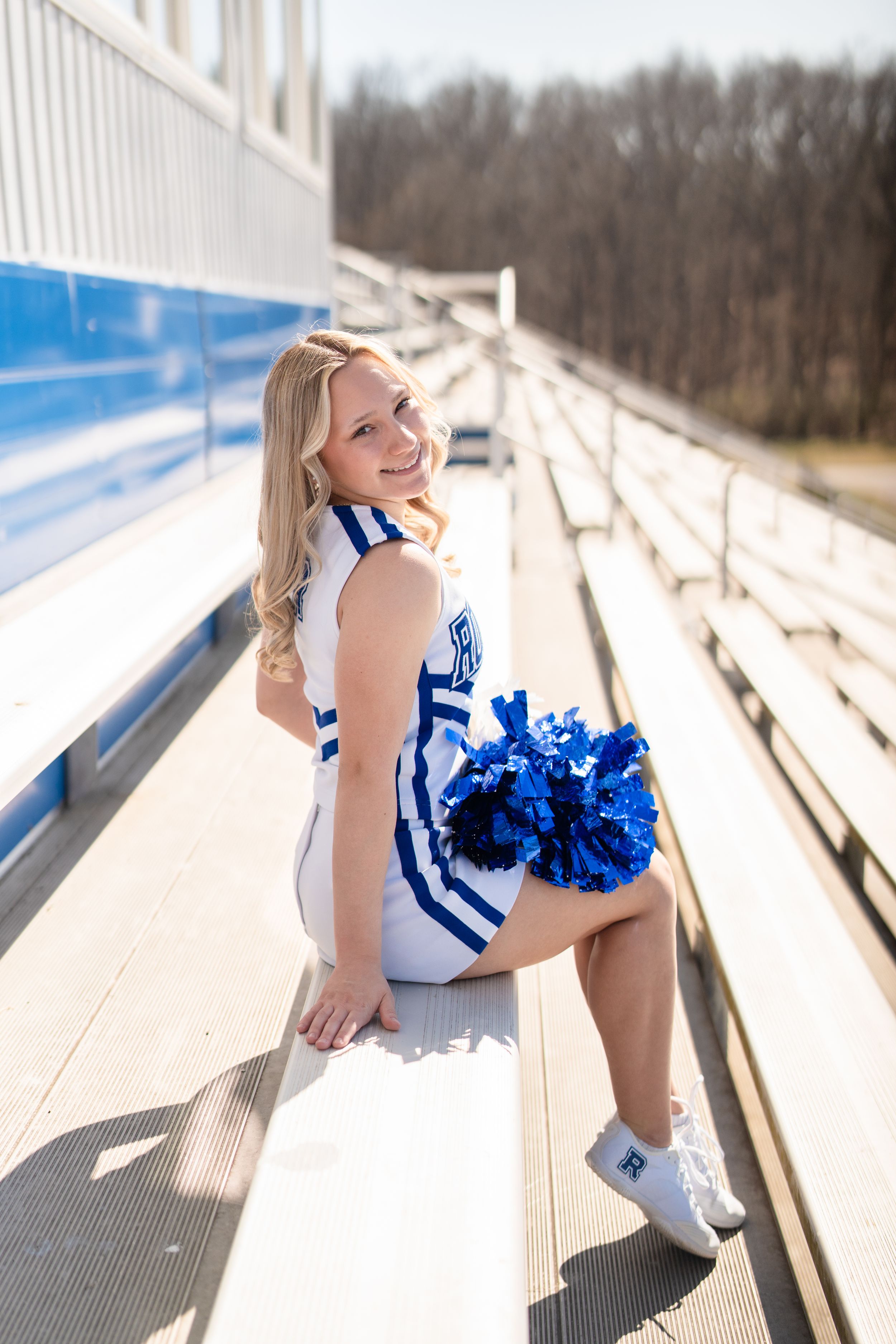 Cheerleader in blue and white uniform posing on bleachers holding blue pom-poms outdoors.