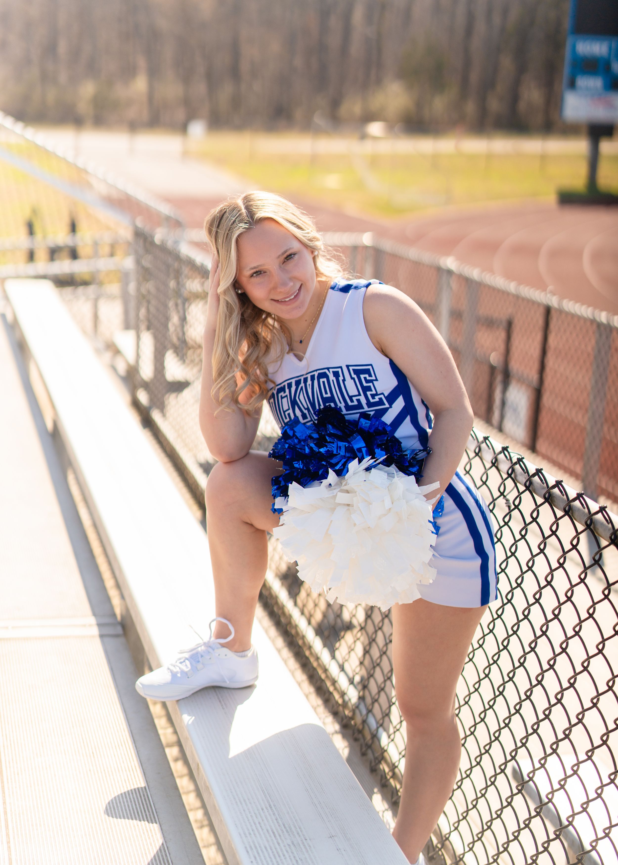 Blonde cheerleader in blue and white uniform leaning on bleacher fence holding white pom-poms.