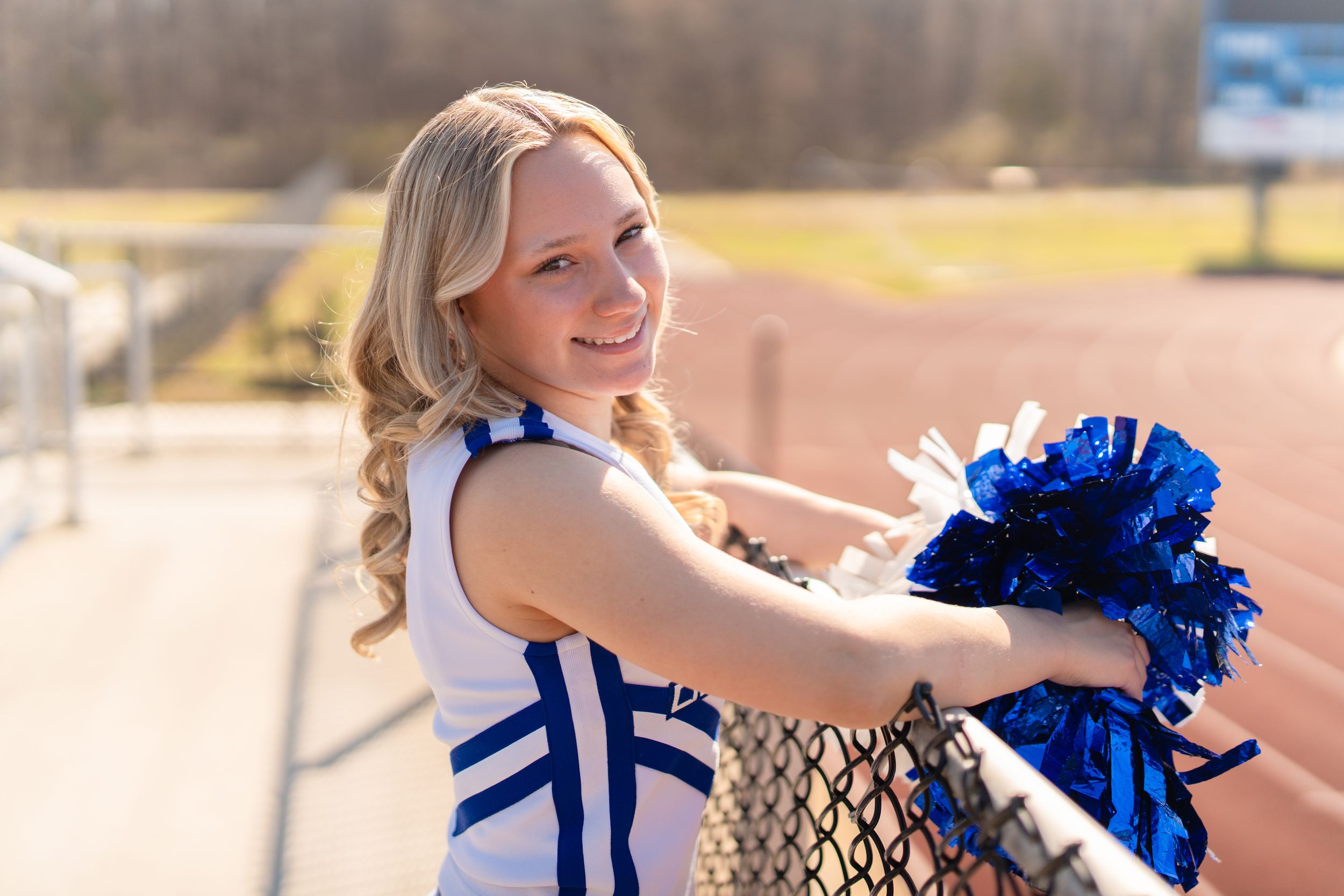 Young cheerleader in blue and white uniform holding blue pom-poms, smiling at a stadium railing.
