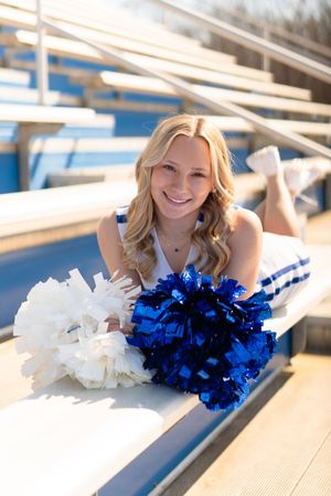 Smiling Rockvale cheerleader lying on bleachers with blue and white pom poms in her school uniform.