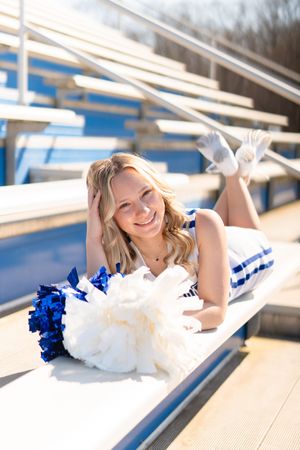 Smiling cheerleader in blue and white uniform lying on bleachers holding pom-poms.