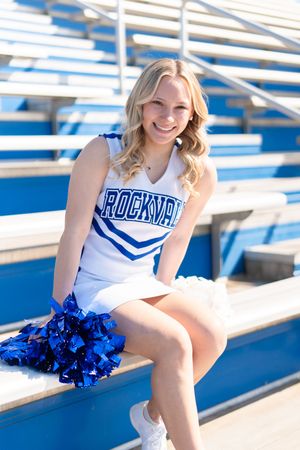 Rockvale cheerleader sitting on stadium bleachers smiling while holding blue pom poms in white uniform.