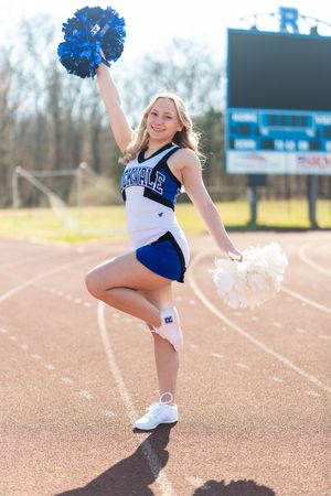 Rockvale cheerleader in blue and white uniform posing on track with one leg raised holding pom poms.