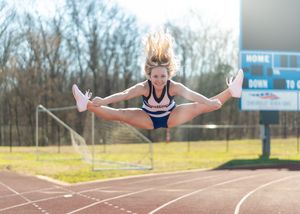 Cheerleader performing a toe-touch jump split in mid-air on a track field with bleachers in background.