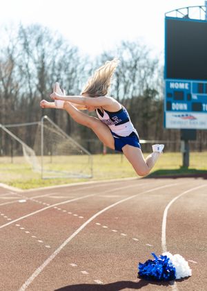 Cheerleader performing a high jump on a track field with pom-poms on ground.