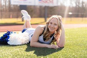Cheerleader in uniform lying on green turf field, smiling with blue and white pom-poms on a sunny day.