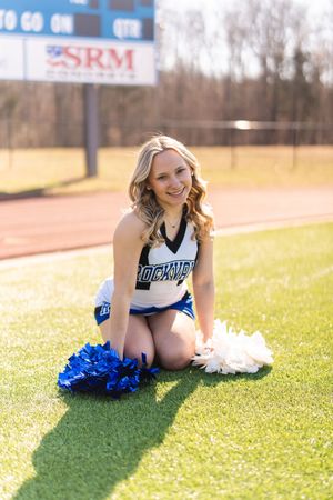 Smiling cheerleader in blue and white uniform kneeling on green turf field holding blue and white pom-poms.