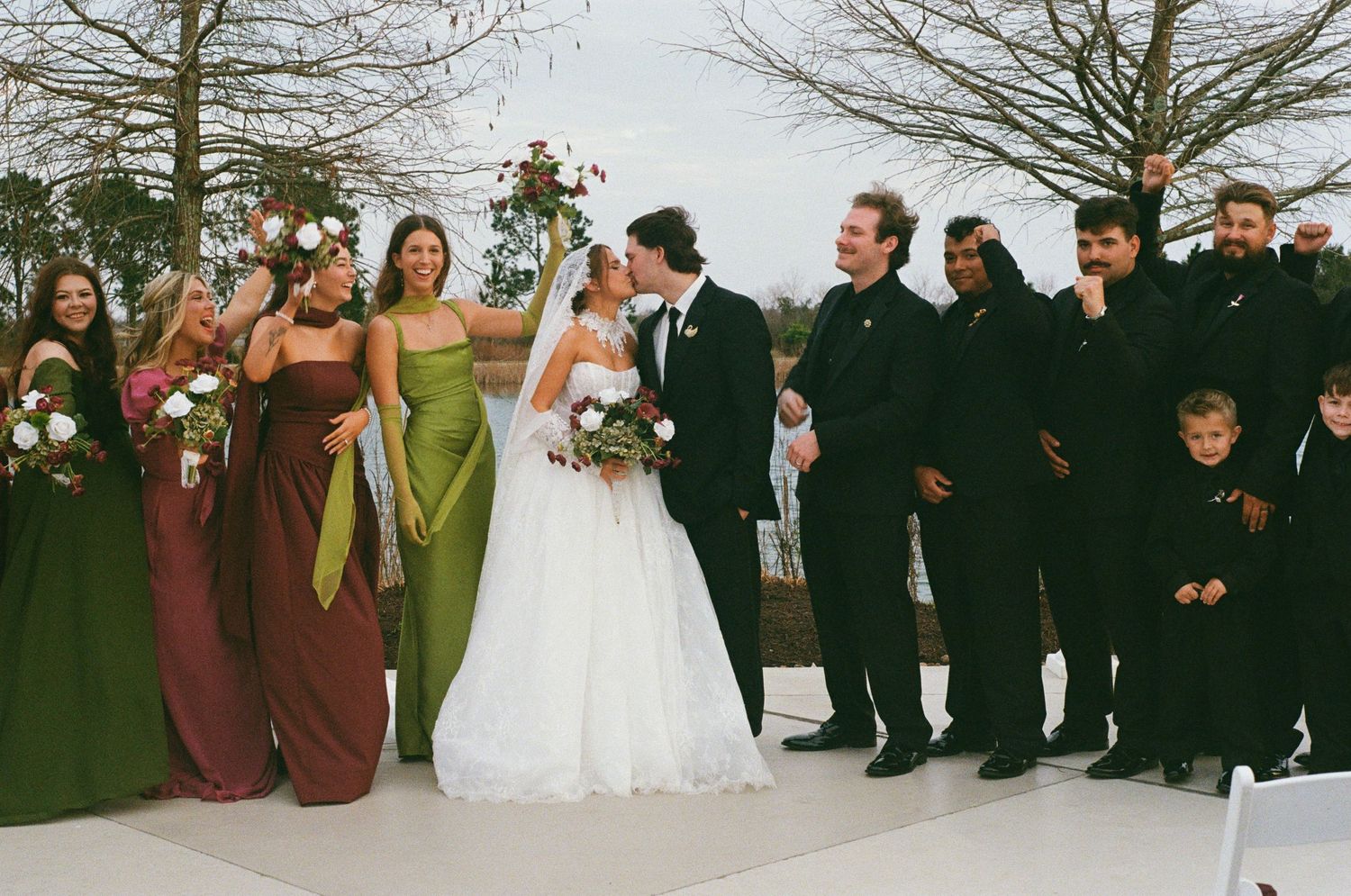Wedding party poses outdoors, bride and groom kissing, bridesmaids in green and brown gowns, groomsmen in black suits.