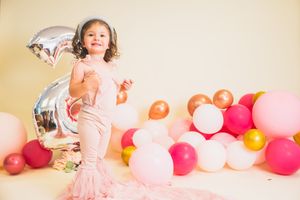 A toddler girl in a pink ruffle outfit poses with colorful balloons and a silver number 2 balloon at her birthday party.