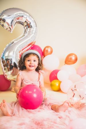 Smiling toddler girl in pink tutu celebrates 2nd birthday with colorful balloons and silver number 2 balloon.