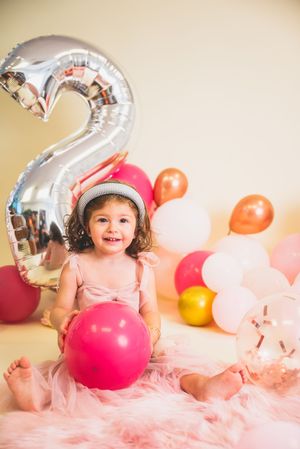 Smiling toddler girl in pink tutu dress celebrating 2nd birthday with colorful balloons and silver number 2 balloon.