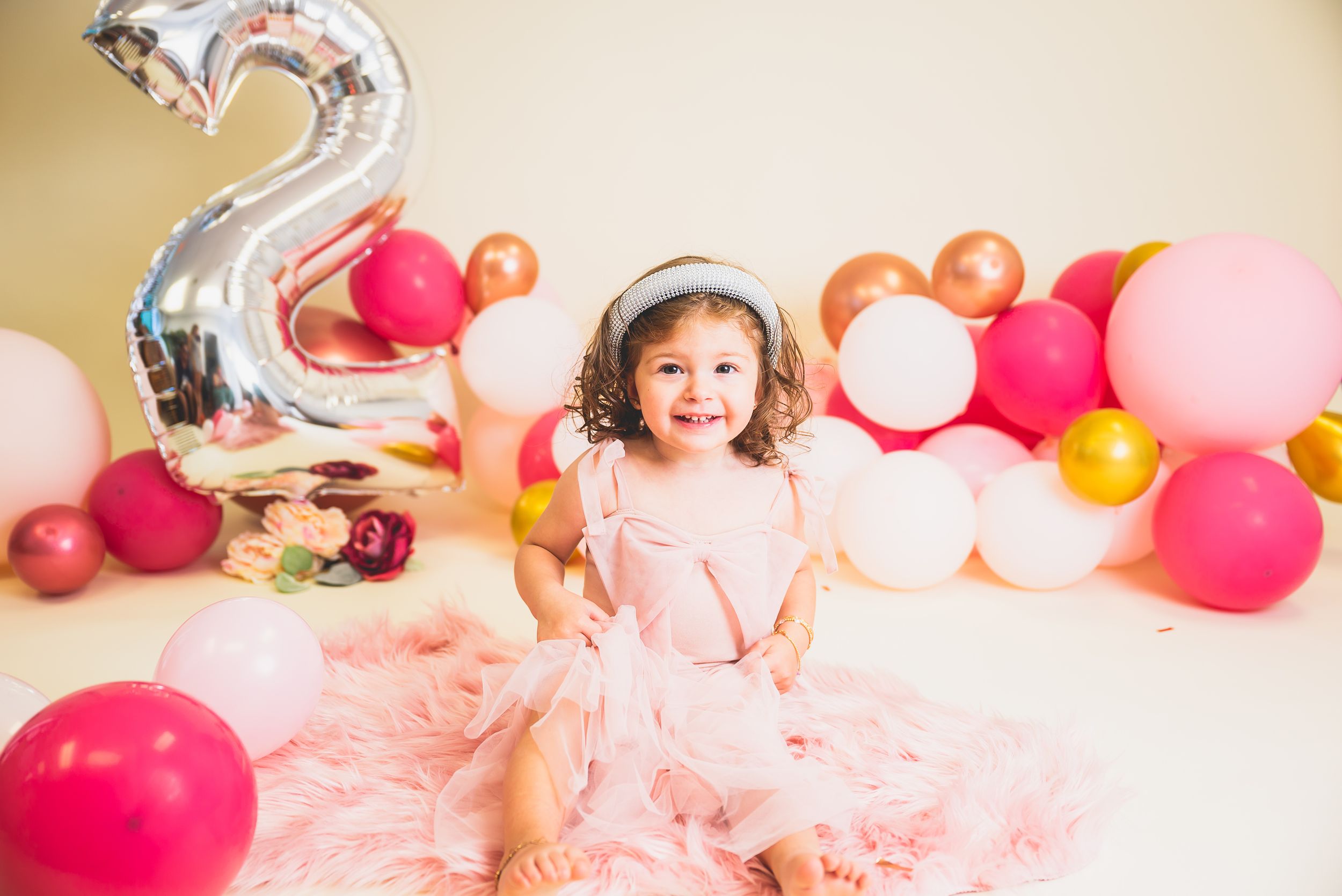 Smiling toddler girl in pink tutu dress sits before colorful balloons and a silver number 2 balloon at her birthday party.