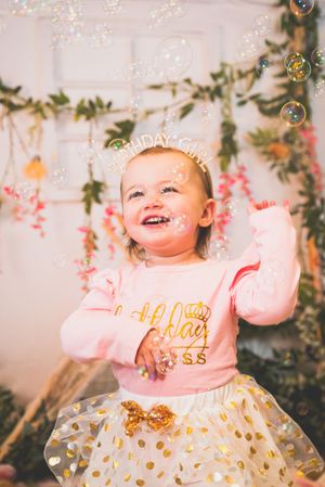 Joyful toddler in pink birthday shirt and gold polka dot skirt laughs while playing with bubbles at her first birthday party.