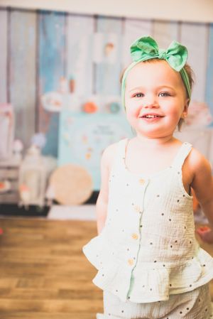 Smiling toddler girl wearing a green bow headband and white dotted outfit, posing happily indoors.