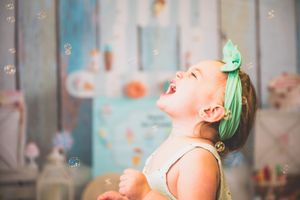 Joyful toddler girl with green bow laughing among floating bubbles in a pastel birthday party setting.