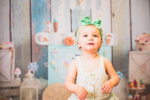 Adorable baby girl in green bow headband and white dress gazing upward against colorful ice cream themed backdrop.