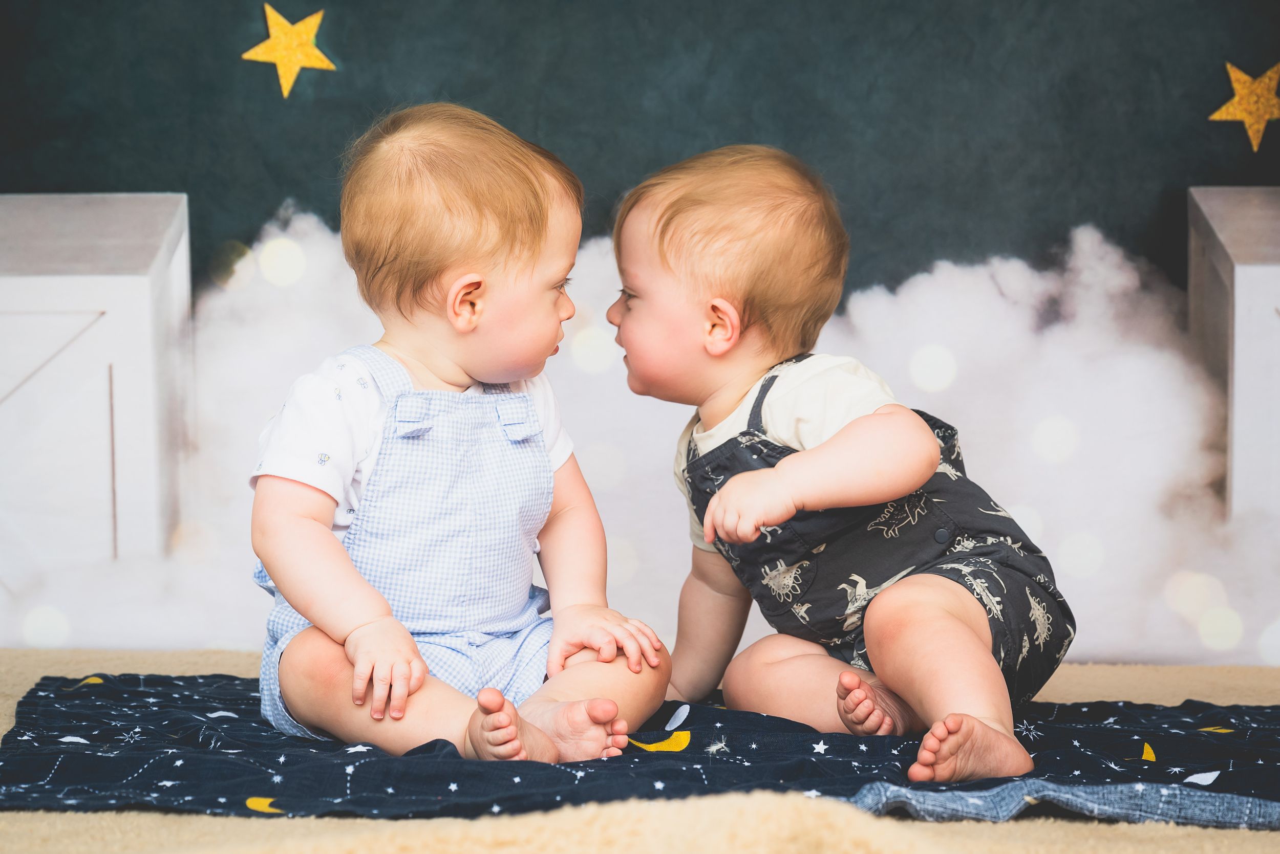 Two adorable babies in cute outfits sitting face to face, appearing to kiss, with star and cloud decorations.